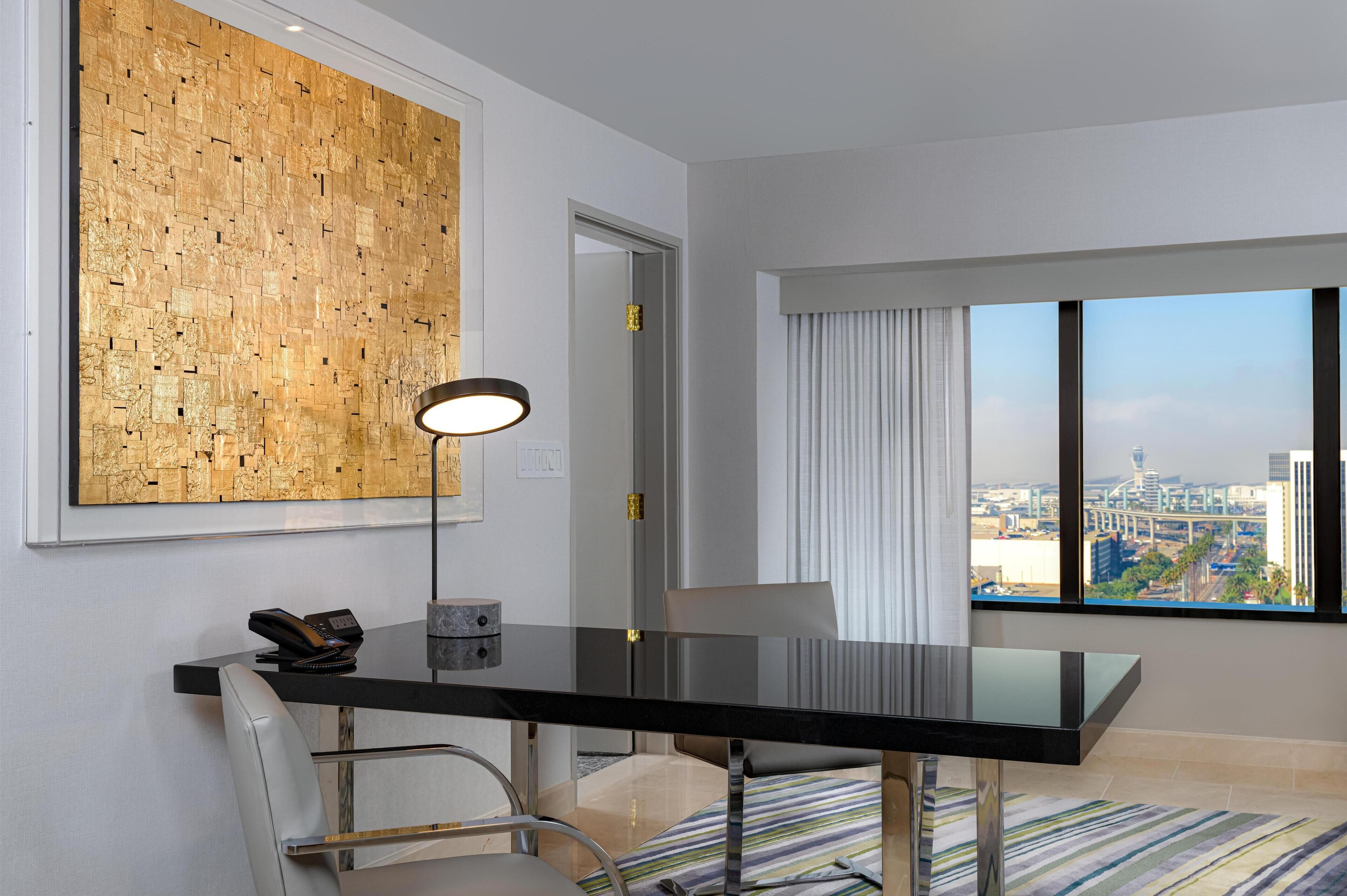 A modern hotel room with a large window offering a city view, a black dining table, and a unique textured wall art piece.