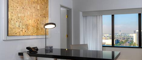 A modern hotel room with a large window offering a city view, a black dining table, and a unique textured wall art piece.