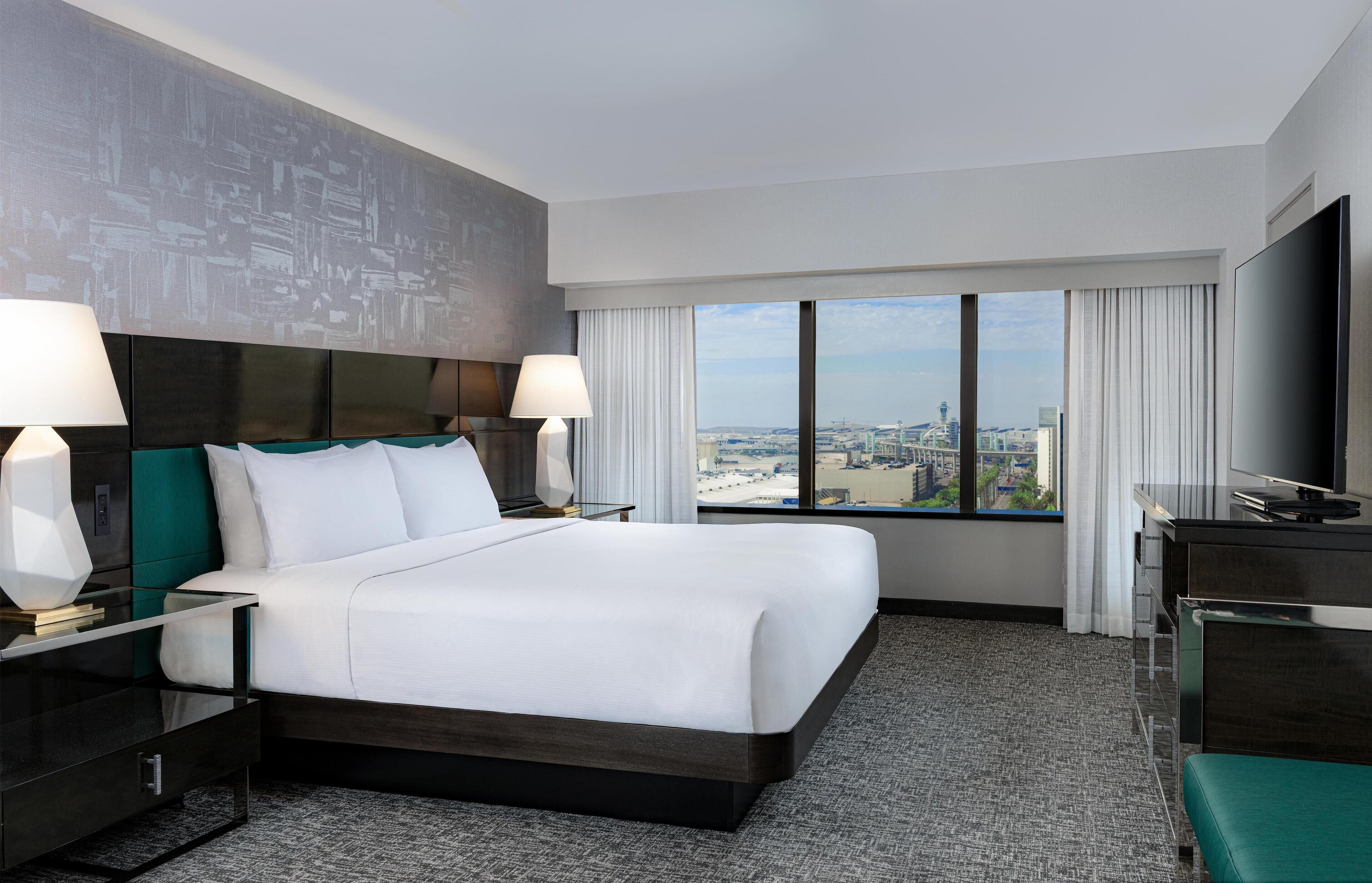 A hotel room with a large bed, two bedside tables with lamps, a desk with a flat-screen TV, and a view of a cityscape through large windows.