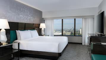 A hotel room with a large bed, two bedside tables with lamps, a desk with a flat-screen TV, and a view of a cityscape through large windows.