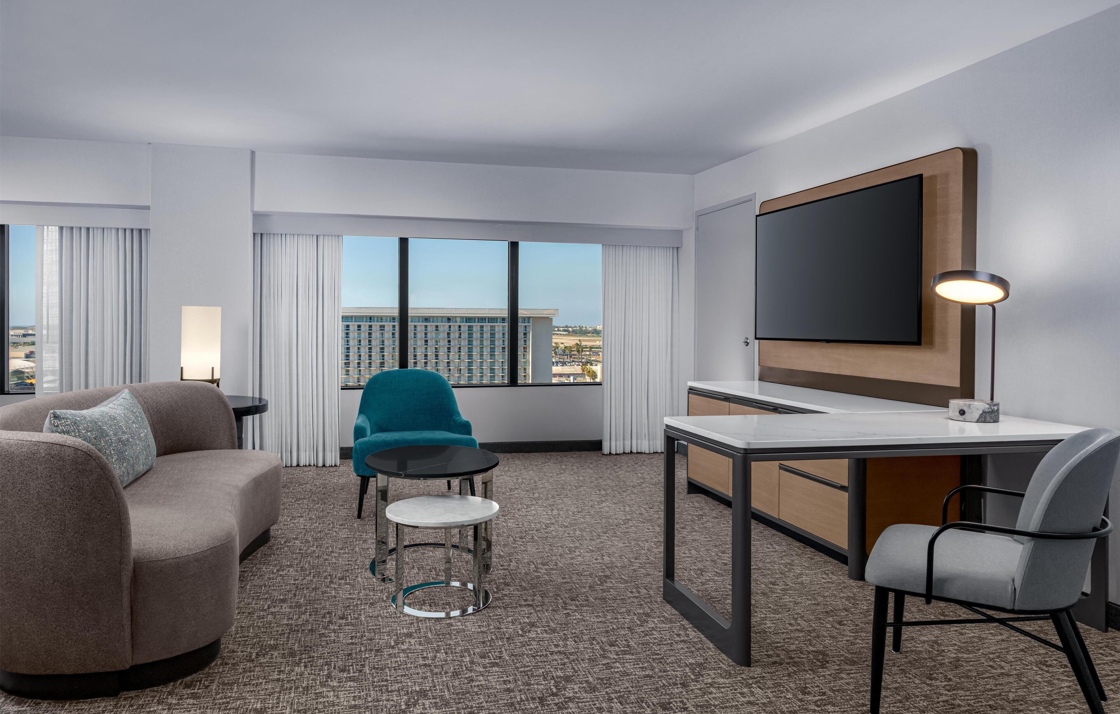 Ein Hotelzimmer mit einer Couch, einem Sessel, einem Schreibtisch, einem Flachbildfernseher und Blick auf eine Stadtlandschaft durch große Fenster.