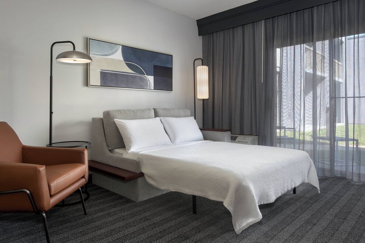 Una habitación de hotel moderna con una cama grande, un sofá, una silla, un escritorio y un ventanal con cortinas.