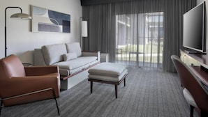 A modern hotel room with a sofa, armchair, ottoman, flat-screen TV, and large windows with sheer curtains. - Courtyard by Marriott Poughkeepsie (Poughkeepsie)