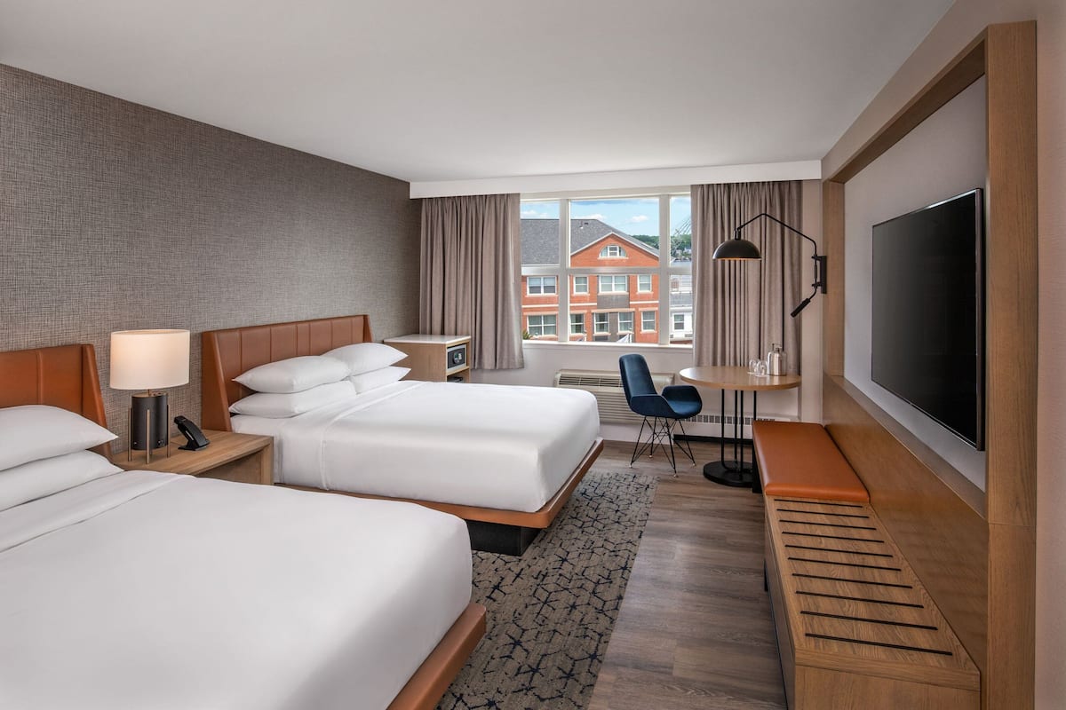 A hotel room with two beds, a desk, a TV, and a view of buildings outside.