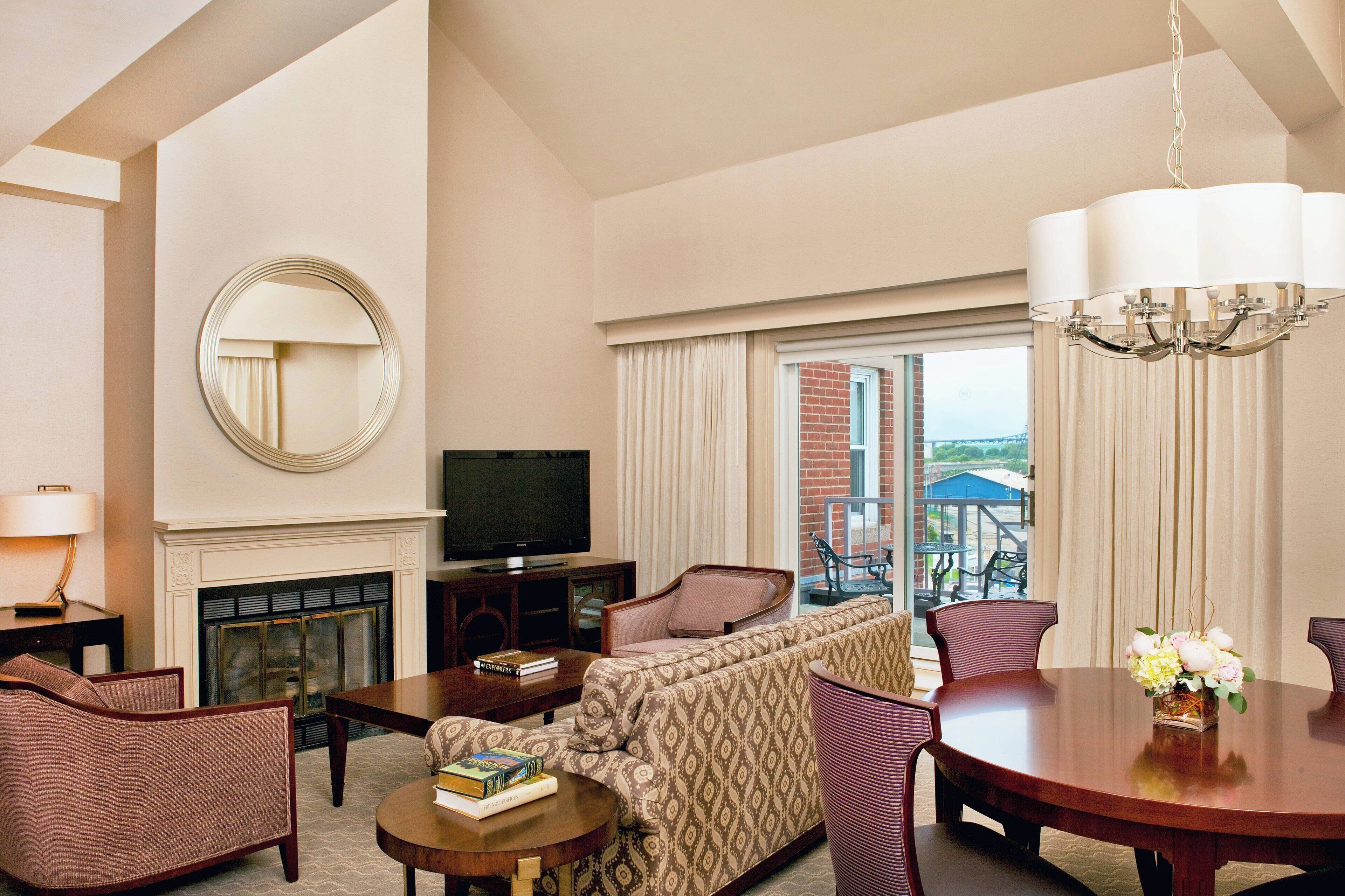 A hotel room with a fireplace, a television, a sofa, a dining table, and a view of the outside.