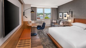 A hotel room with a large bed, a desk with a chair, a TV, a window with a view of a body of water and buildings, and a sofa with a lamp. - Sheraton Portsmouth Harborside Hotel (Portsmouth)