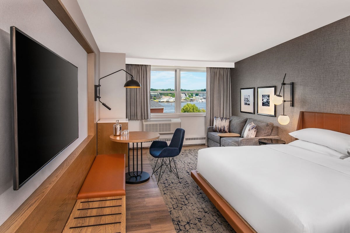 A hotel room with a large bed, a desk with a chair, a TV, a window with a view of a body of water and buildings, and a sofa with a lamp.