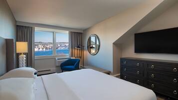 A hotel room with a large bed, a desk, a chair, a dresser, a flat-screen TV, a window with a view of the city, and a lamp.