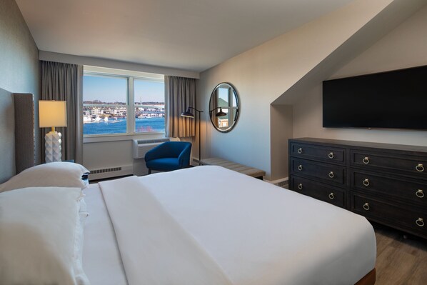 A hotel room with a large bed, a desk, a chair, a dresser, a flat-screen TV, a window with a view of the city, and a lamp.
