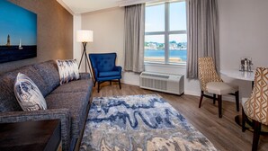 A hotel room with a large window, a sofa, a blue armchair, a dining table with chairs, and a patterned rug. - Sheraton Portsmouth Harborside Hotel (Portsmouth)