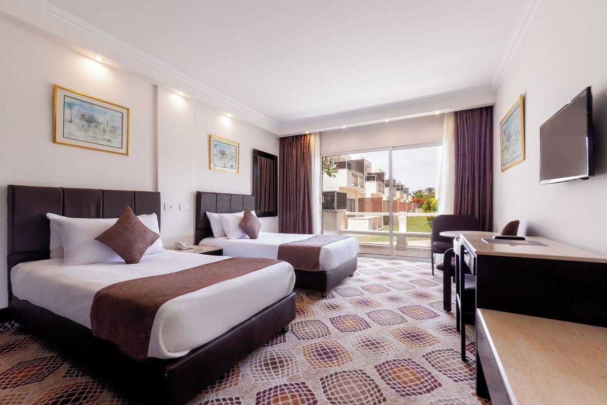 A hotel room with two beds, a desk, a chair, and a large window with a view of buildings.