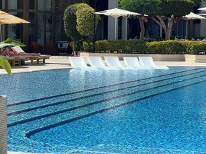 A swimming pool with lounge chairs and umbrellas, surrounded by greenery and a building facade.