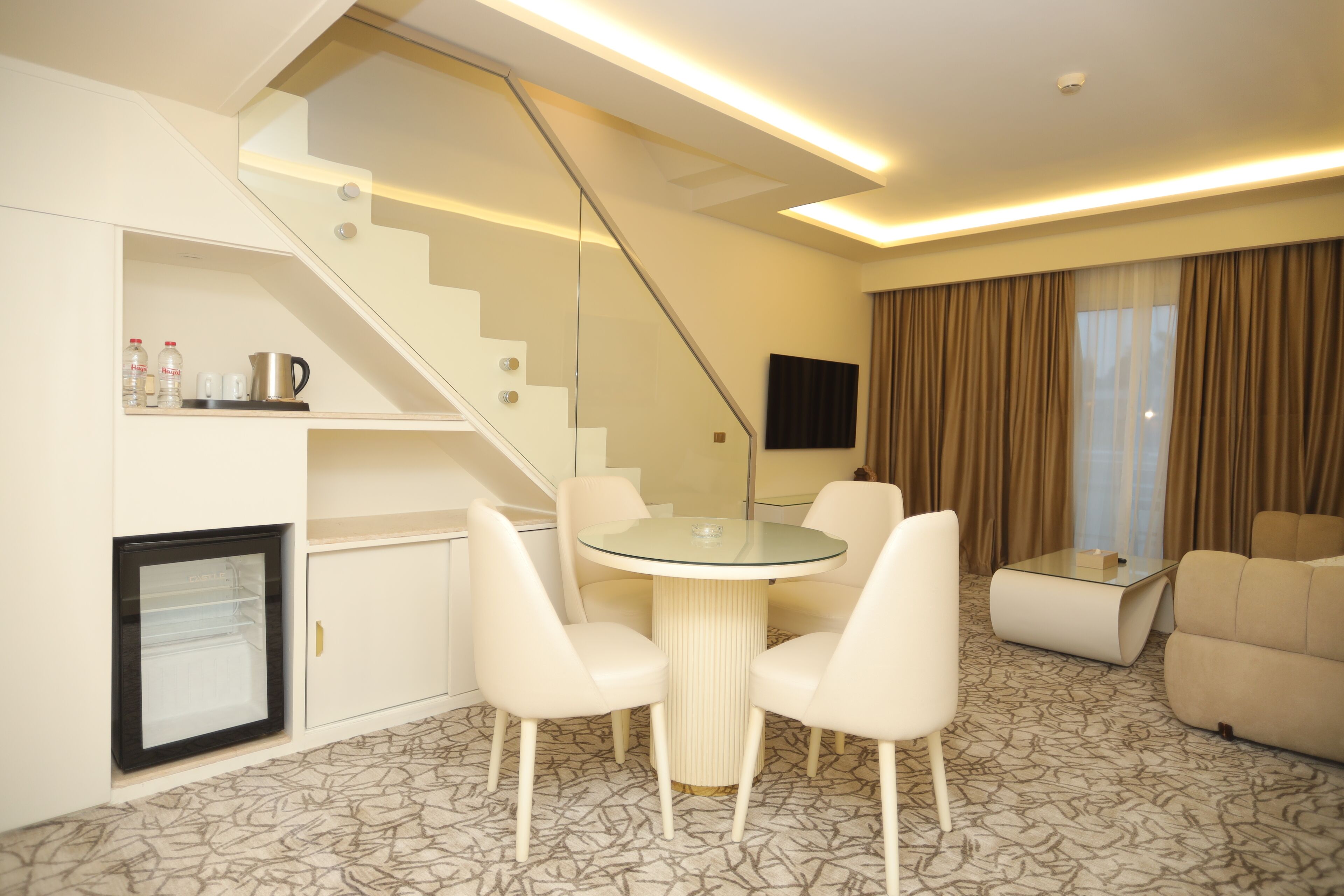 A modern hotel room with a dining area, a sofa, a flat-screen TV, and a staircase.
