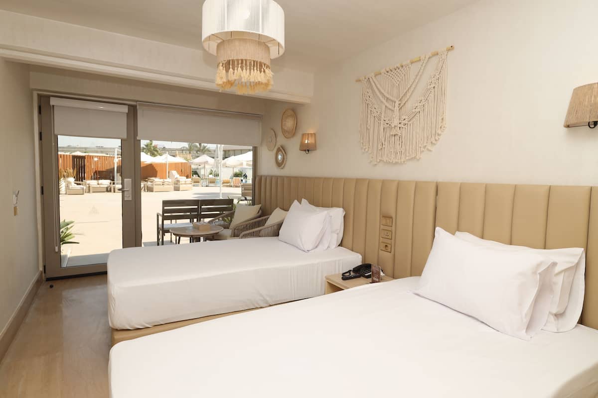 A hotel room with two beds, a chandelier, macrame wall decor, and a view of the outdoor area.