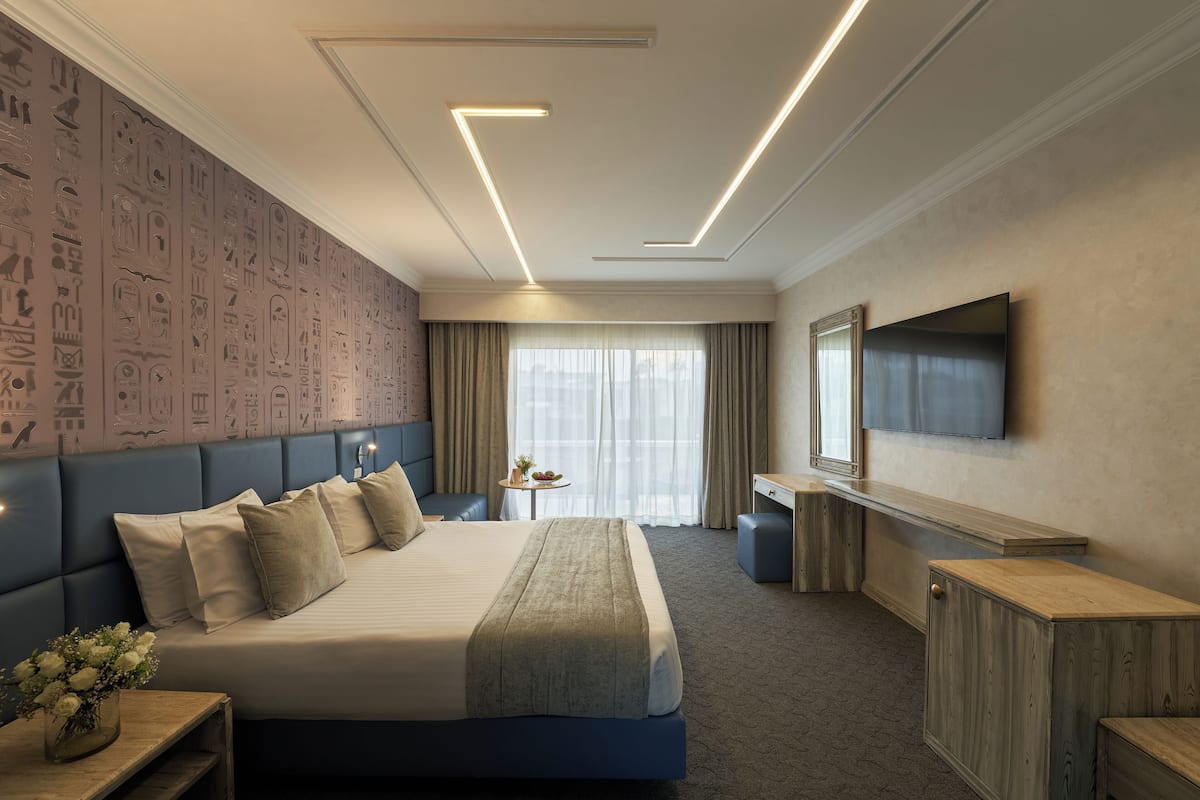 A modern hotel room with a large bed, a flat-screen TV, a desk, and a seating area with a view of the city.