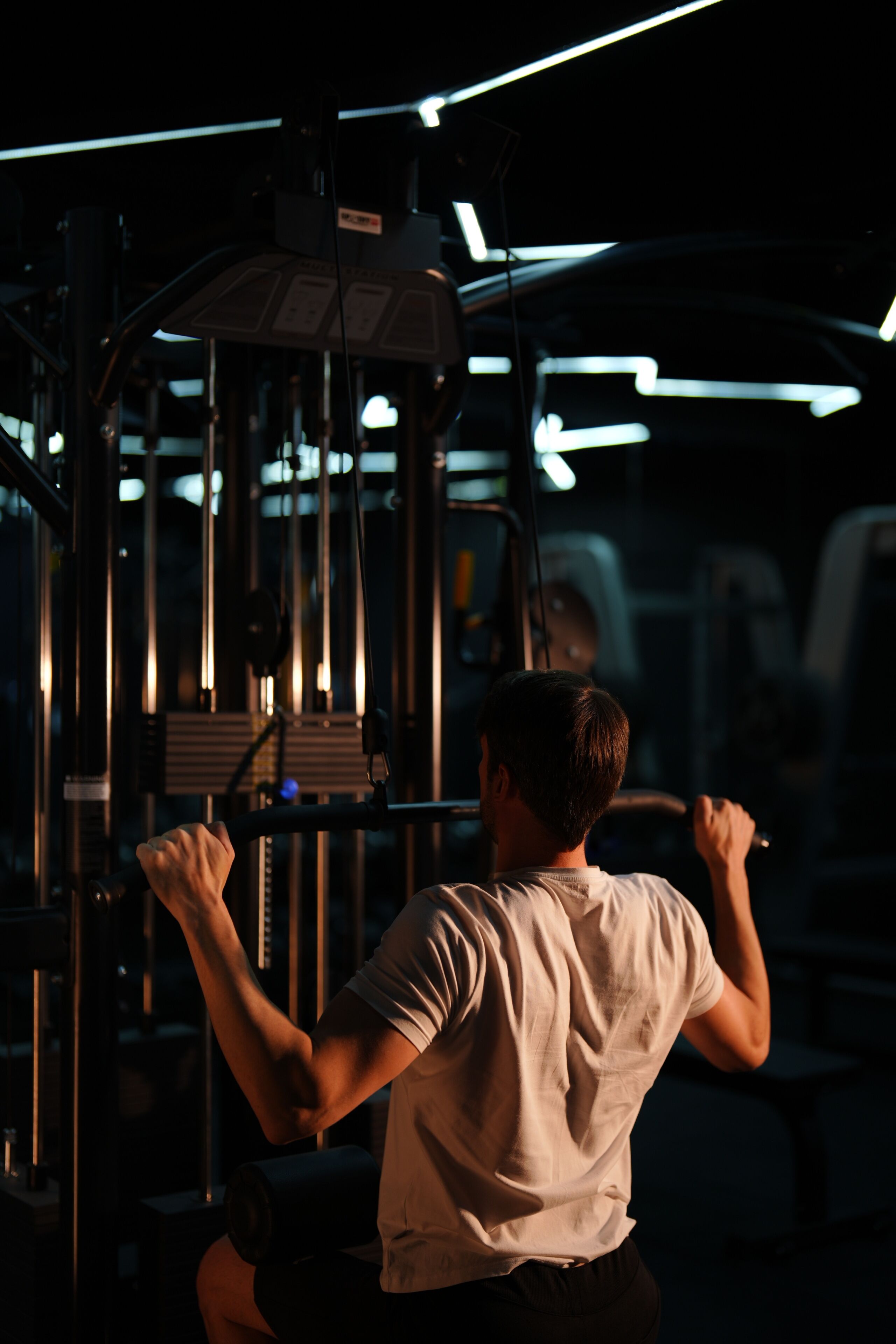 Una persona usando una máquina de poleas en un gimnasio, con equipo visible e iluminación en el techo.