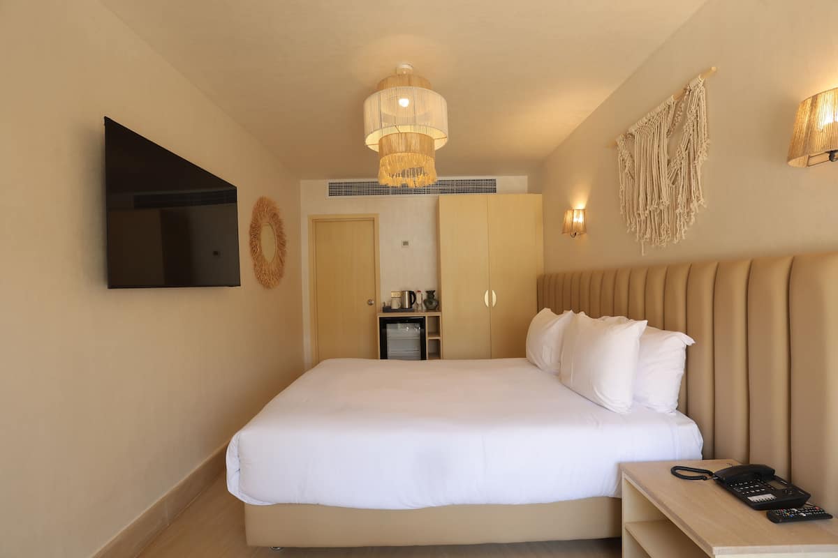 A hotel room with a bed, bedside table, lamp, wall-mounted TV, and a small kitchenette area.