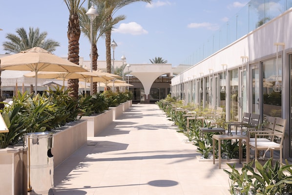 A pathway lined with outdoor seating, palm trees, and a modern building with large windows. - Pyramids Park Resort Cairo (Giza)