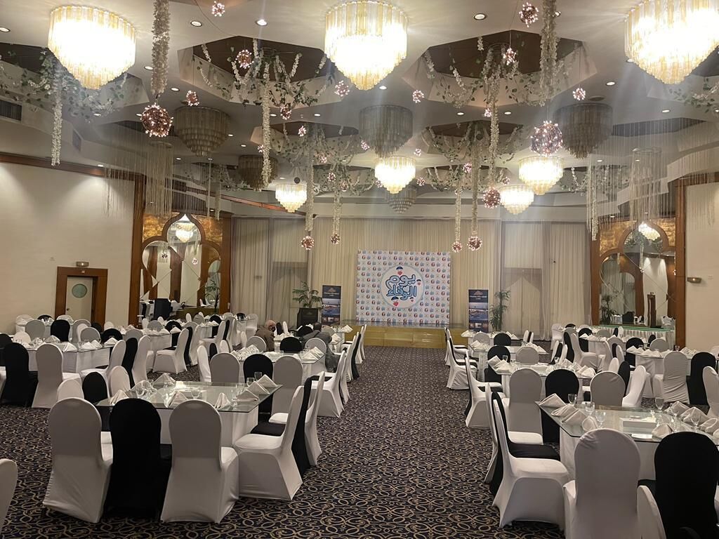 Un salón de banquetes con mesas redondas, sillas blancas, candelabros y un escenario con fondo decorativo.