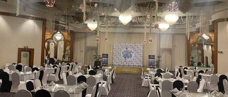 Un salón de banquetes con mesas redondas, sillas blancas, candelabros y un escenario con fondo decorativo.