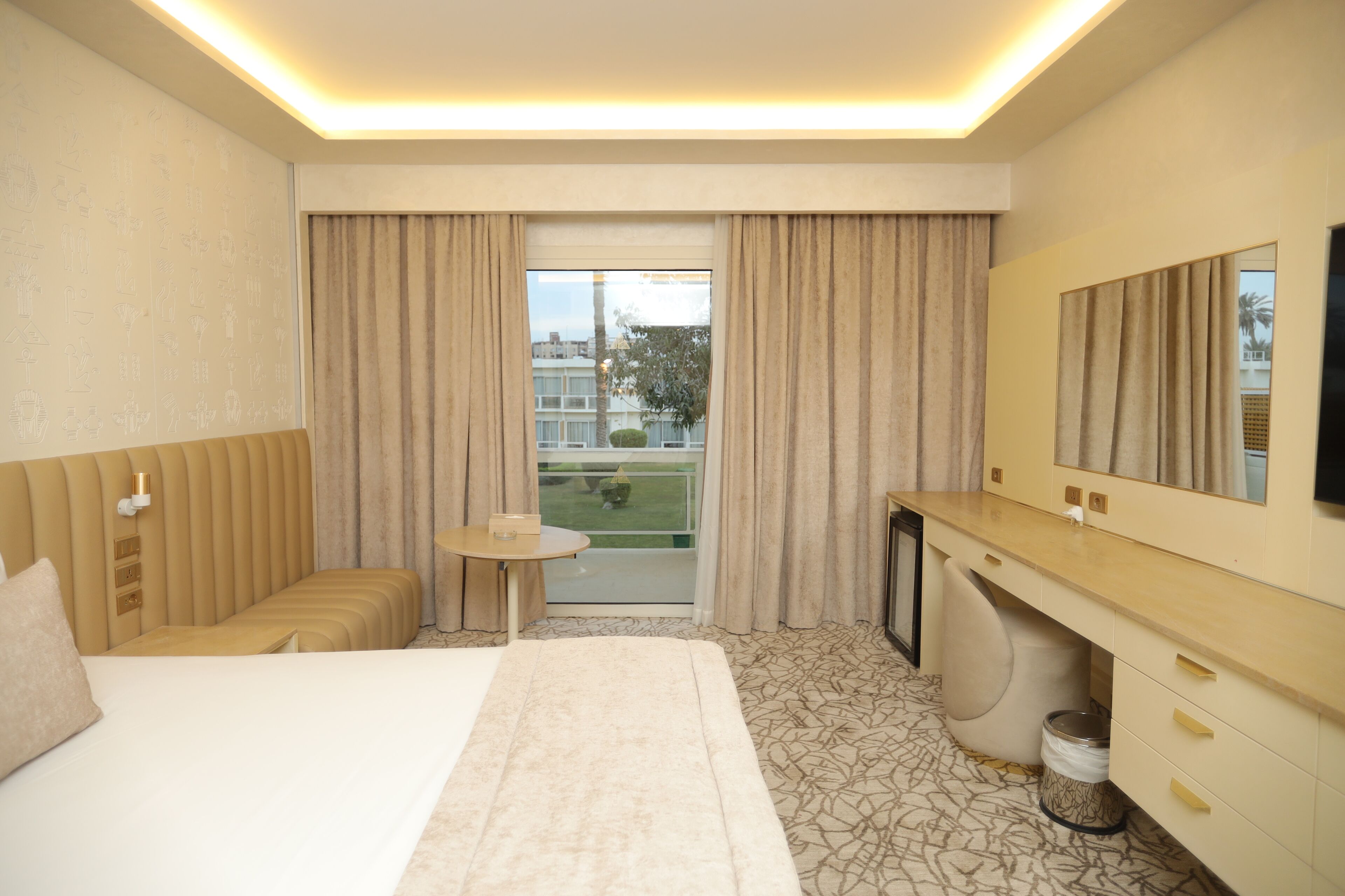 A hotel room with a bed, a desk, a chair, a small table, a mirror, and a view of the outside.