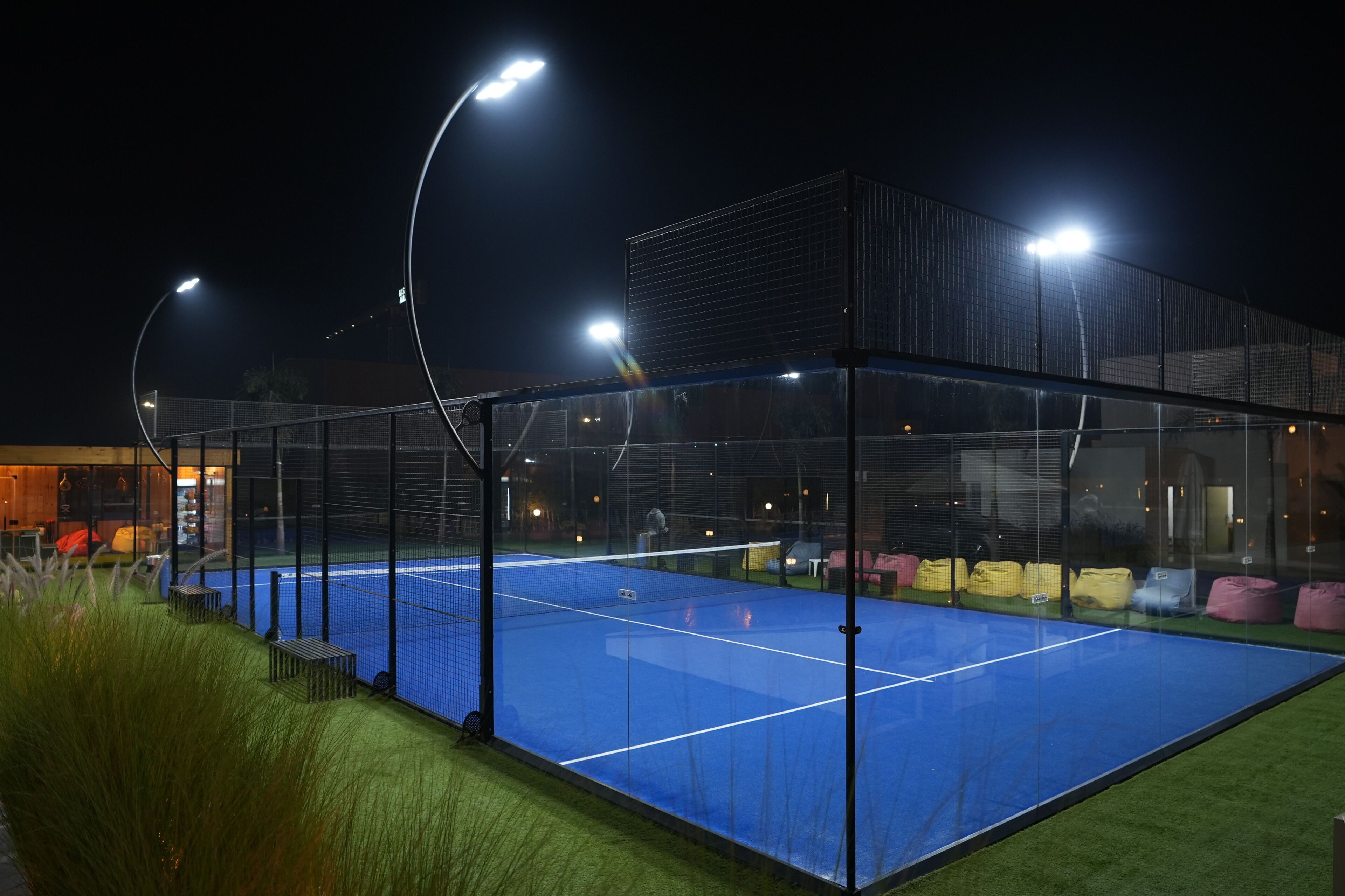 A tennis court with a net, surrounded by a fence, and illuminated by overhead lights.