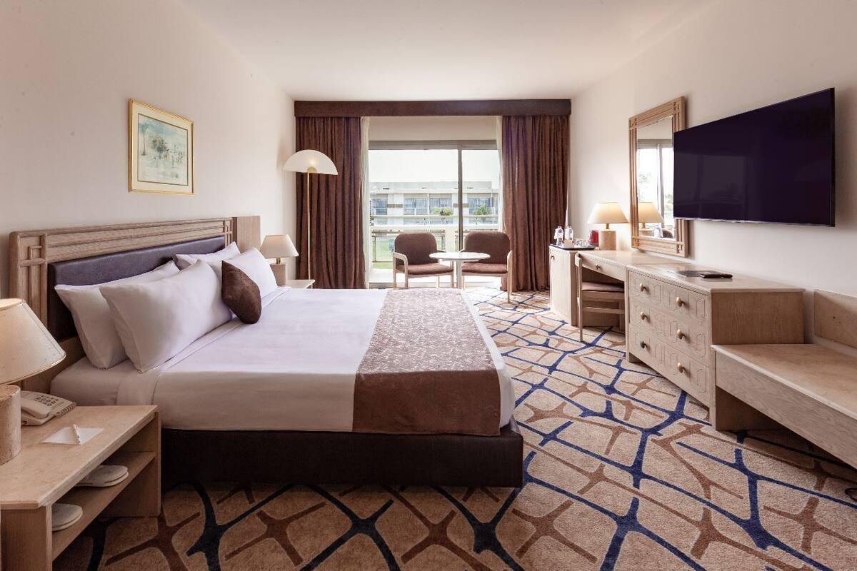 A hotel room with a large bed, a desk, a chair, a TV, and a balcony with a view.