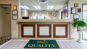 A reception area with a green 'Quality' mat, framed certificates, a clock, and a potted plant. - Quality Inn Oakwood - Gainesville (Flowery Branch)