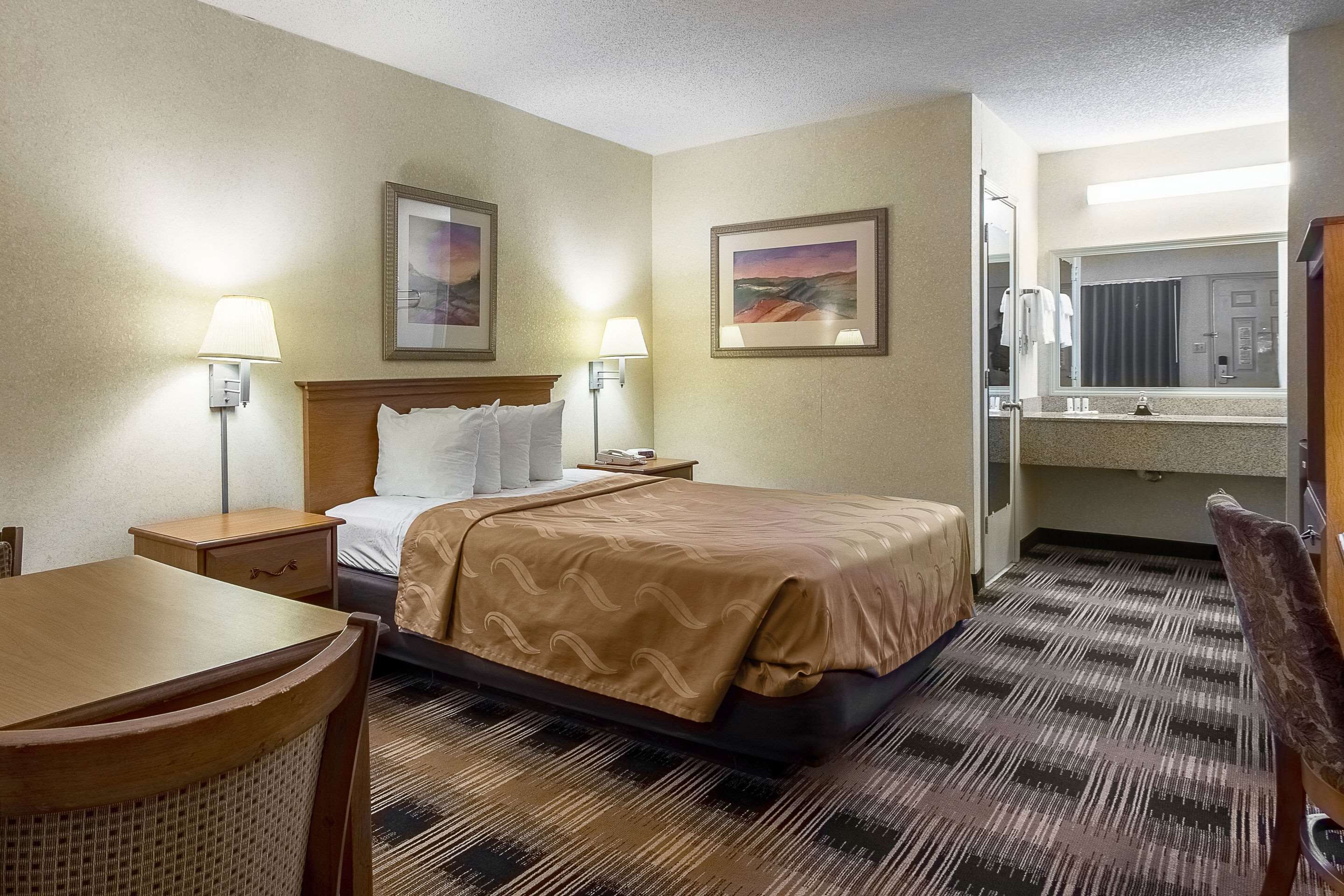 A hotel room with a bed, bedside lamps, a desk, a chair, a bathroom mirror, and two framed pictures on the wall.