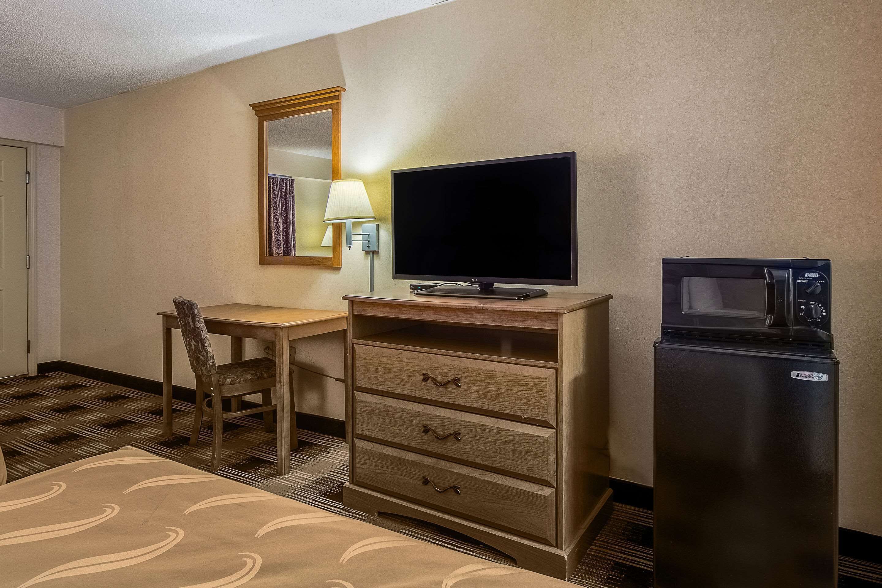 Ein Hotelzimmer mit einem Bett, einer Holzkommode mit Schubladen, einem Fernseher, einer Mikrowelle, einem Stuhl und einem Spiegel.