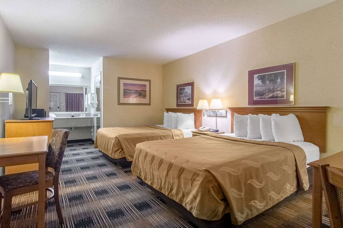 A hotel room with two beds, a desk, a chair, a lamp, and framed pictures on the wall.