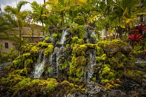 Une cascade avec des rochers couverts de mousse, entourée de plantes tropicales et d’un bâtiment doté d’un toit en tuiles.