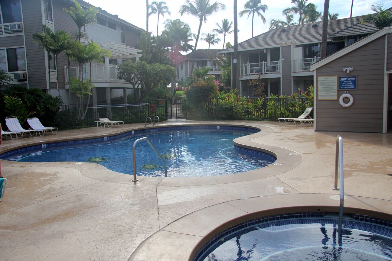 A swimming pool area with lounge chairs and a hot tub, surrounded by buildings and palm trees.