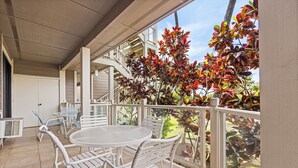 A covered patio area with a round table and white metal chairs, overlooking a garden with red and green foliage. - Wailea Grand Champions - Maui Condo & Home (Kihei)
