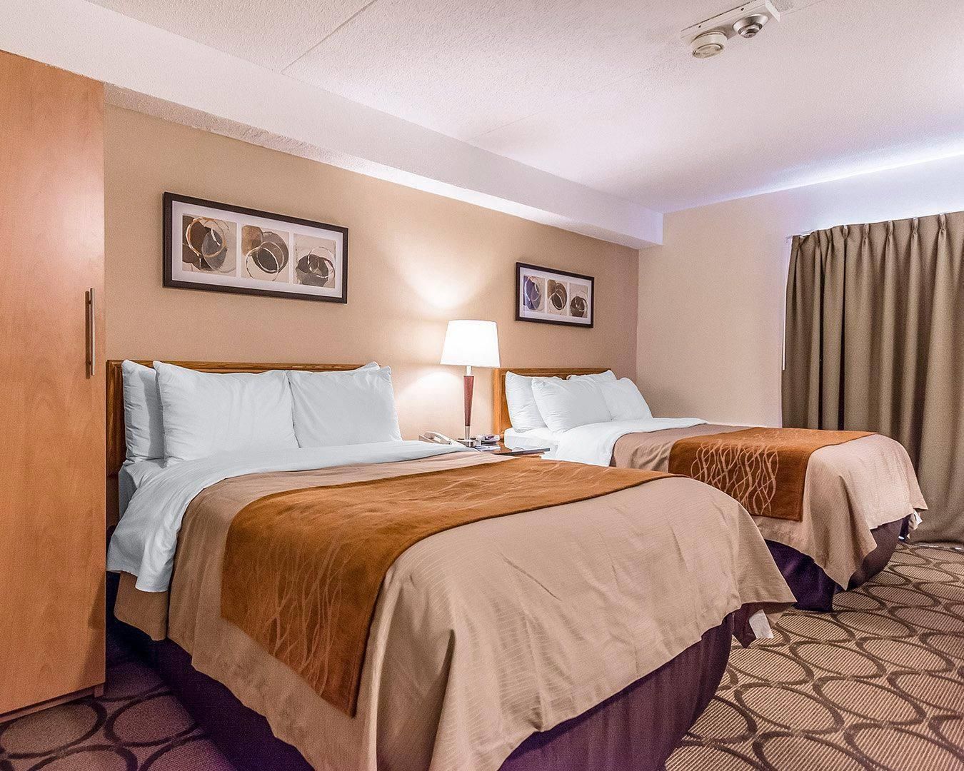 A hotel room with two beds, a nightstand, a lamp, and framed artwork on the wall.