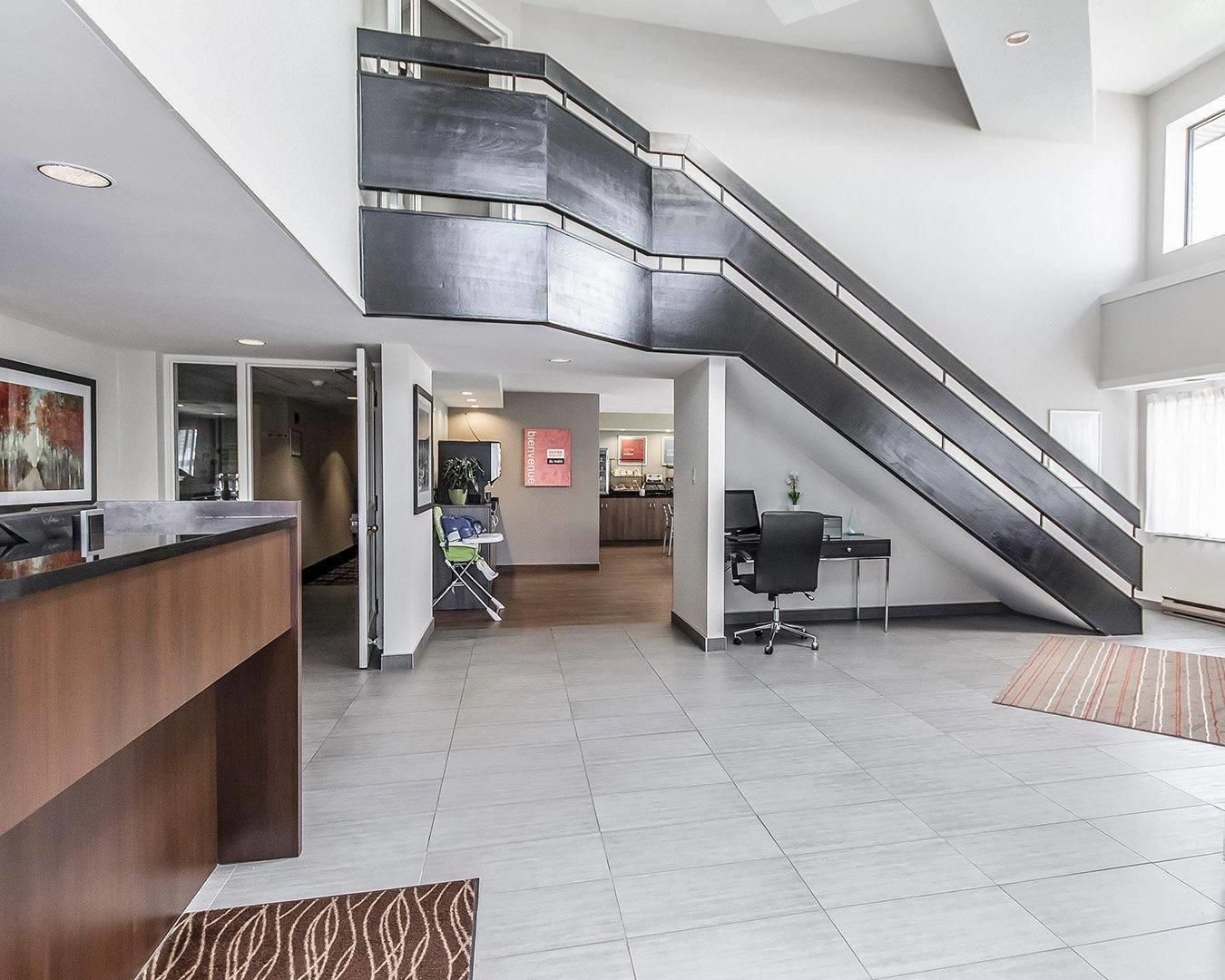 A modern hotel lobby with a central reception desk, a staircase, and a seating area.