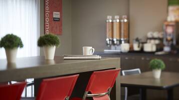 A modern hotel lobby with a reception desk, red chairs, a coffee machine, and a welcome sign.