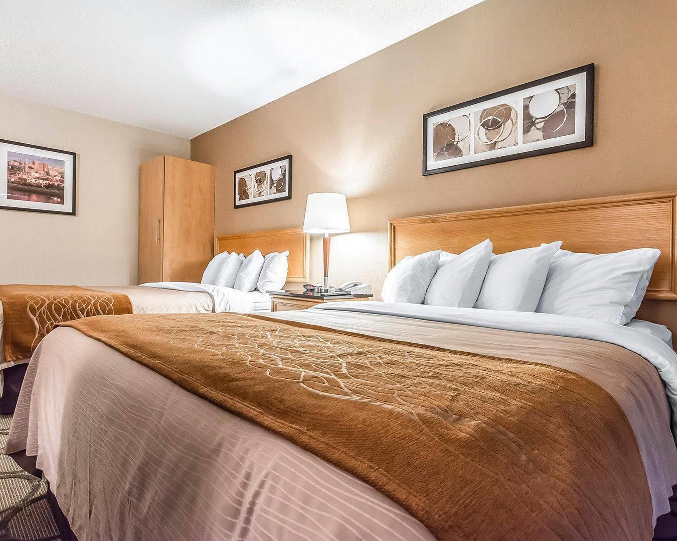 A hotel room with two beds, a wooden headboard, a nightstand with a lamp, and framed artwork on the walls.