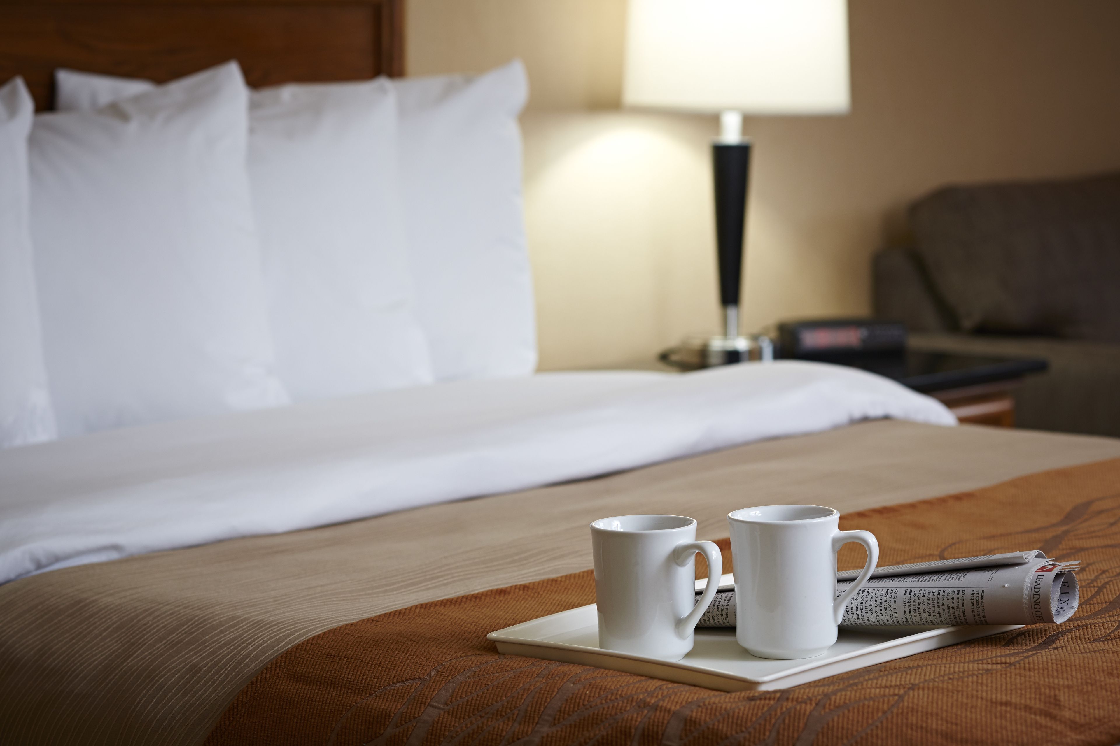 Un lit bien fait, avec du linge de lit blanc, deux tasses blanches posées sur un plateau et un journal roulé sur un coussin marron.