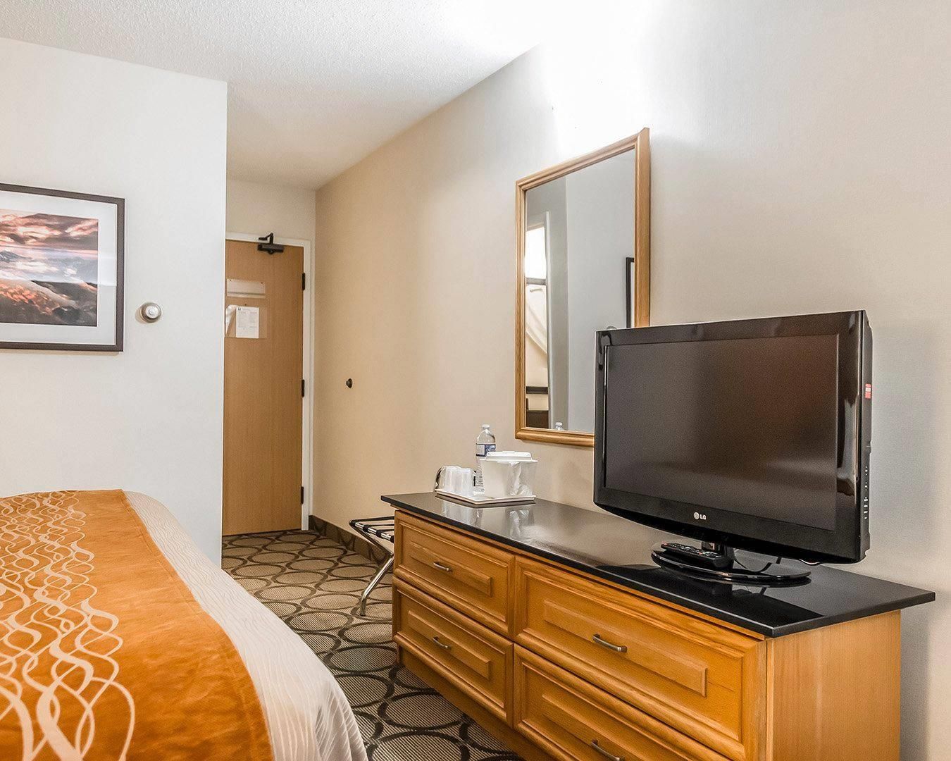 A hotel room with a bed, a television on a wooden dresser, a mirror, and a door leading to a bathroom.
