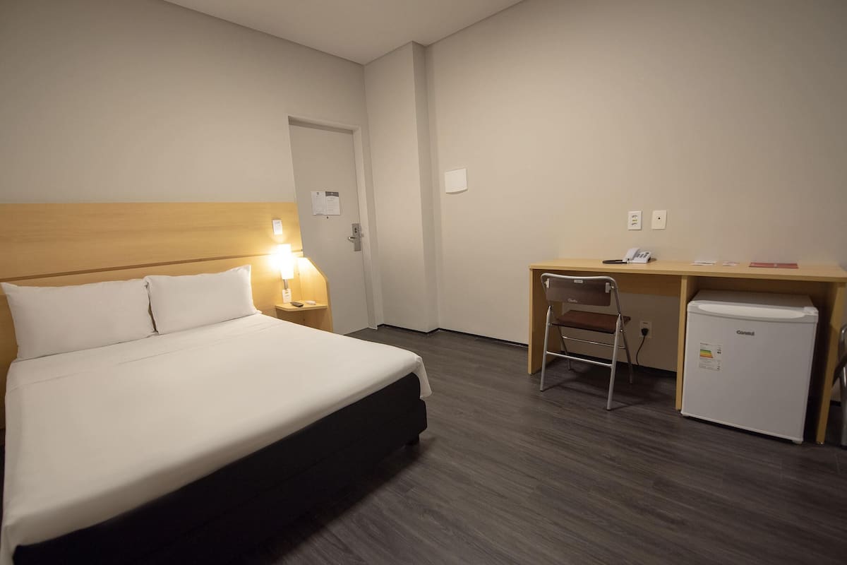 A hotel room with a bed, desk, chair, and a small refrigerator.