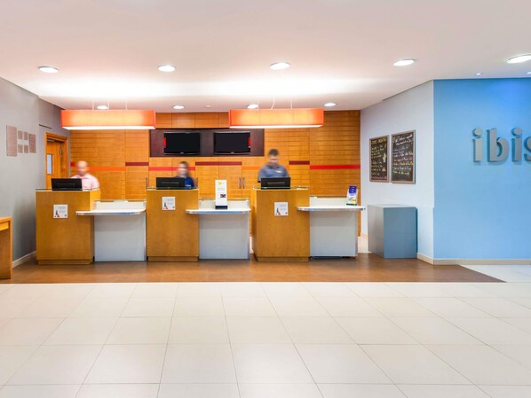 A reception area with four counters, a blue wall with the word 'ibis' on it, and a flat-screen TV mounted above the counters.