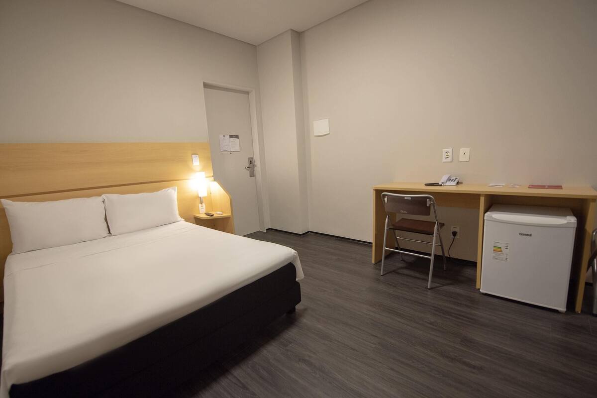 Una habitación de hotel con cama, escritorio, silla y un refrigerador pequeño.