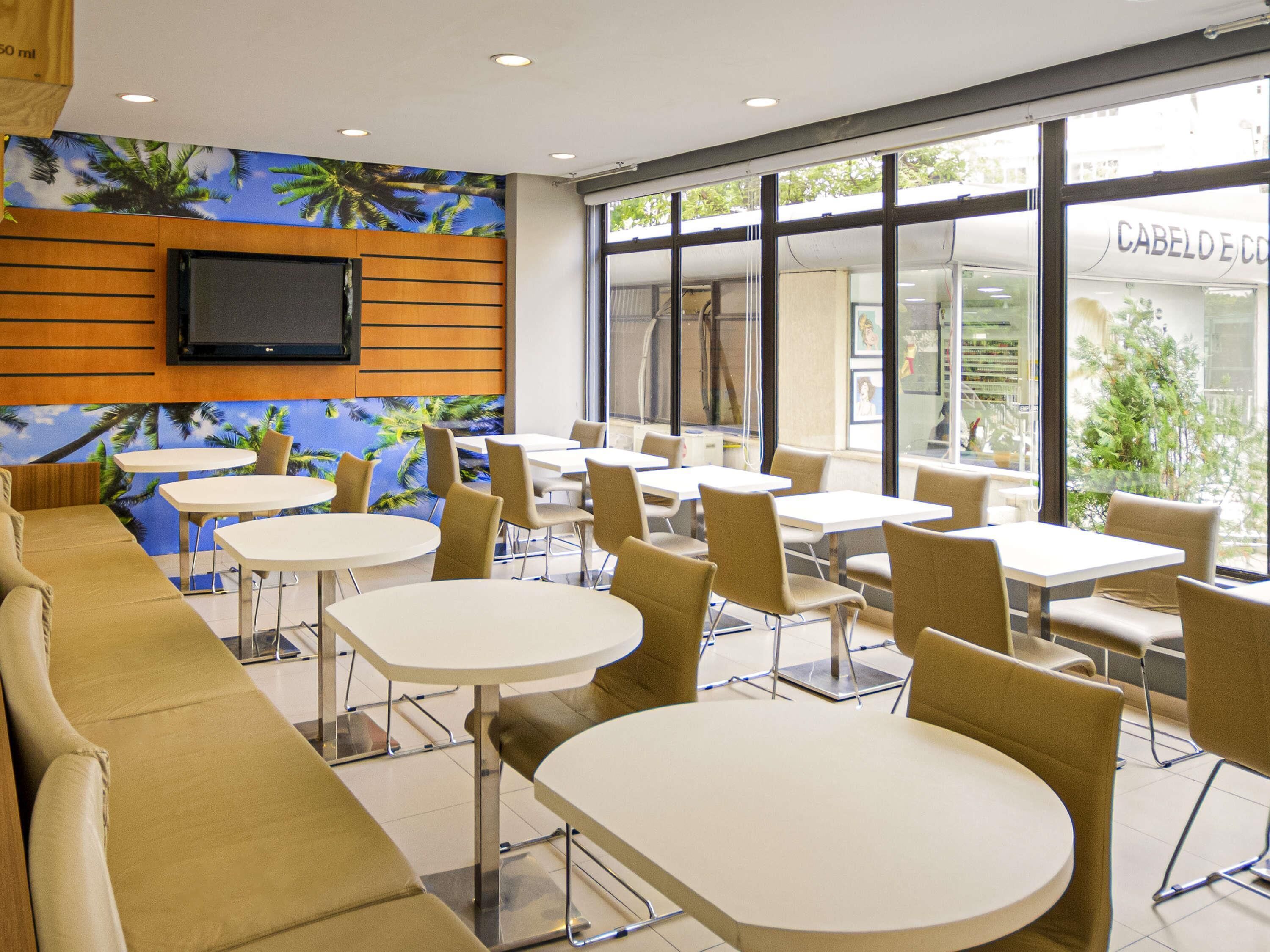 A modern dining area with tables and chairs, a television mounted on the wall, and large windows with a view of the outside.