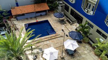 A rooftop pool area with a blue pool, surrounded by potted plants and umbrellas, and a building with a blue facade in the background.