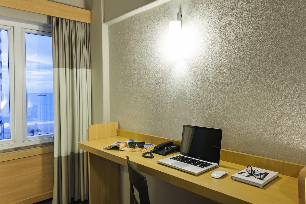 A hotel room with a wooden desk, a laptop, a telephone, a pair of glasses, and a book.