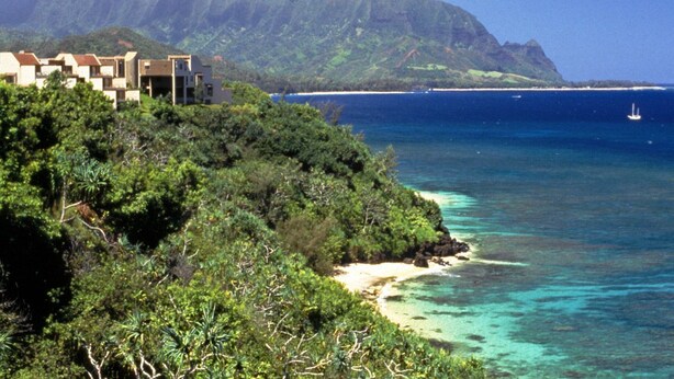 A coastal area with a beach, a building with multiple levels, and a mountainous backdrop. - Castle at Princeville (Princeville)