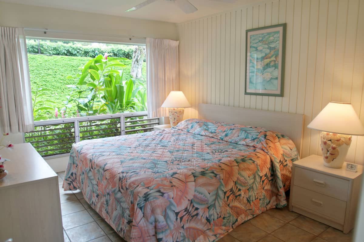 A bedroom with a bed, two bedside tables with lamps, a window with curtains, and a framed picture on the wall.