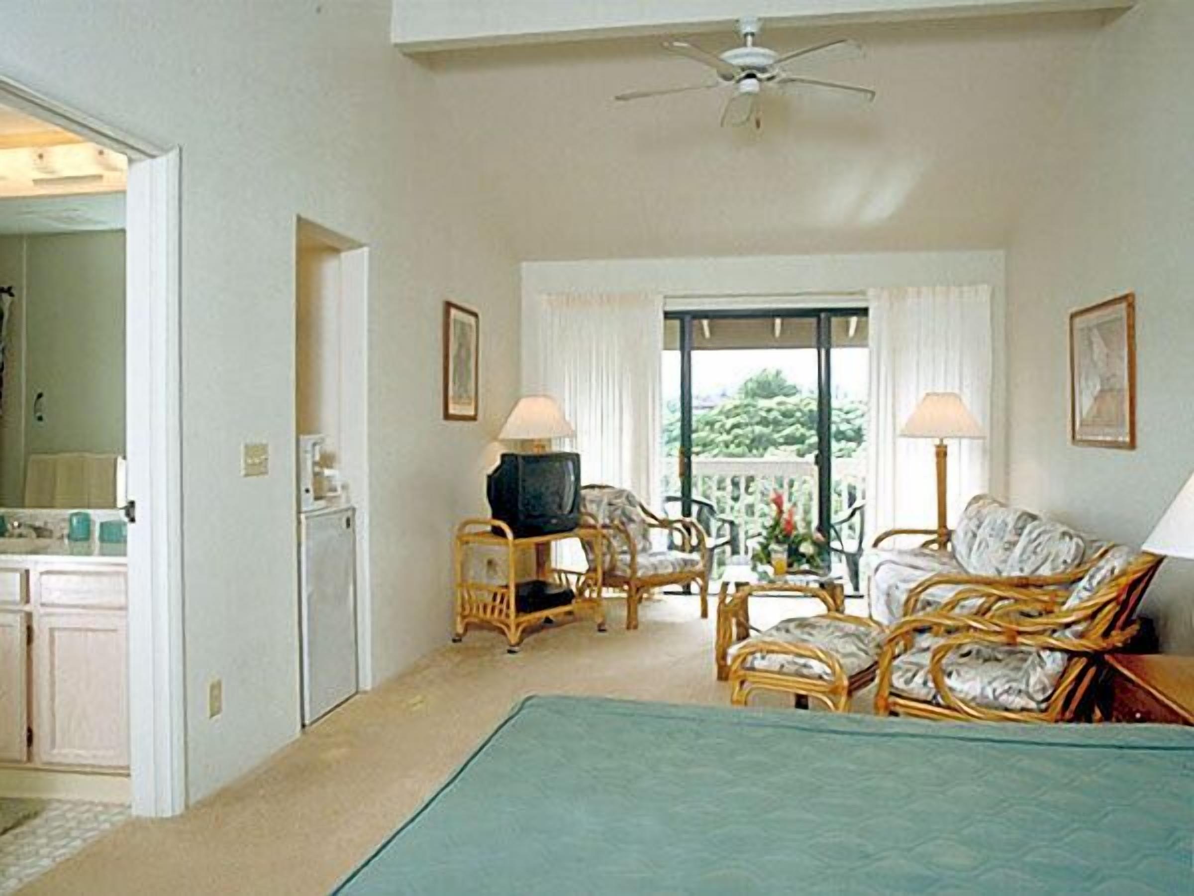 A hotel room with a bed, two wicker chairs, a small table with a lamp, a television, and a balcony with a view of greenery.