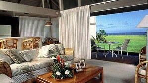 A spacious living room with a large sofa, a coffee table with a floral arrangement, and a view of a grassy area through a sliding door. - Castle at Princeville (Princeville)